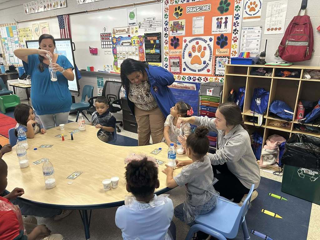 Dr. Andrea Berry-Brown, Superintendent of Schools and a district staff member are assisting young students who are sitting around a table engaging in a classroom experiment. The classroom is featuring educational posters and cubbies.