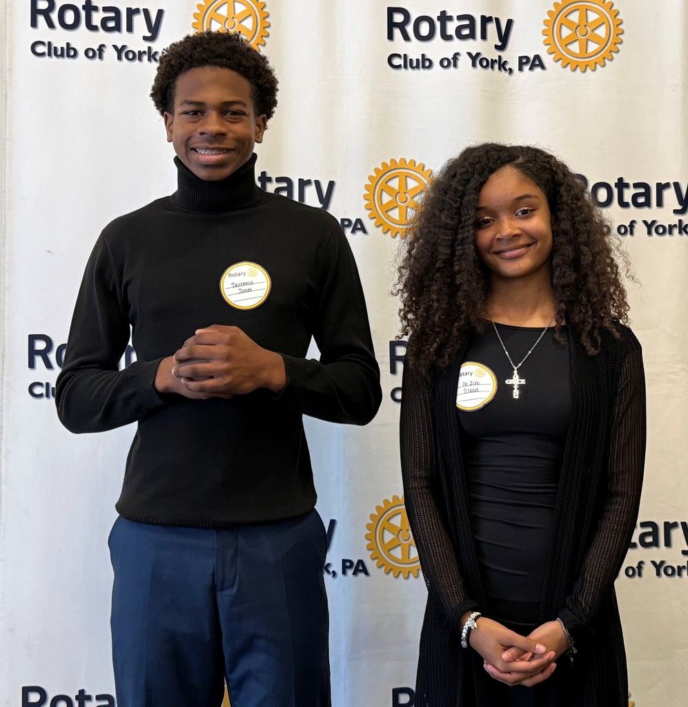 Two William Penn Senior High School students are standing in front of a Rotary Club of York, PA banner, displaying Rotary Club name tags.