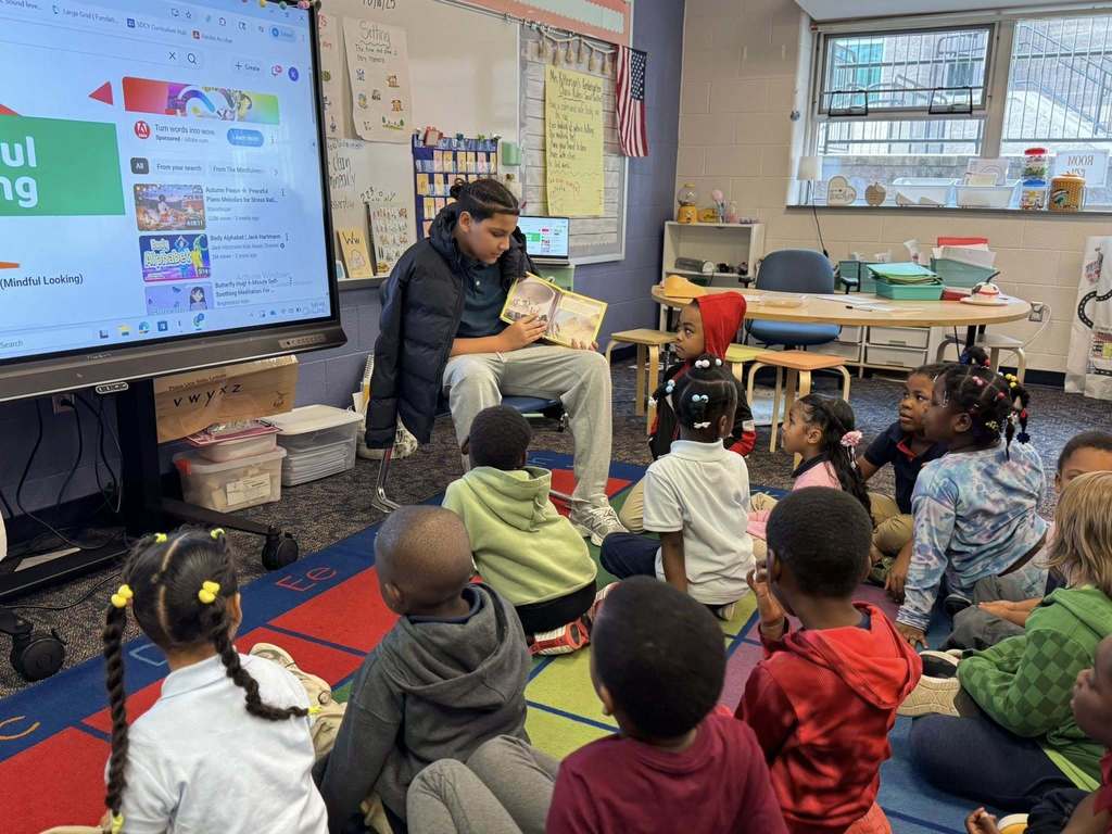 An older student is reading a storybook to a group of younger students who are seated on a colorful carpet in a classroom. 