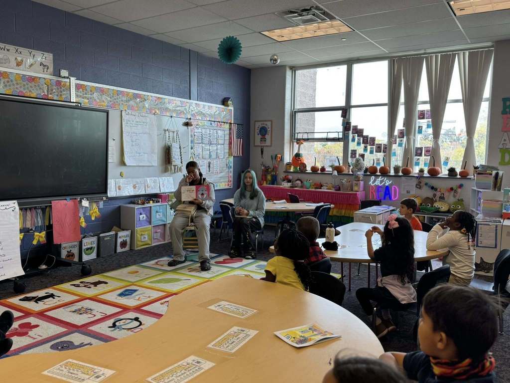 Two older students are reading a storybook to a group of younger students who are seated at a table and on a colorful carpet in a classroom. 
