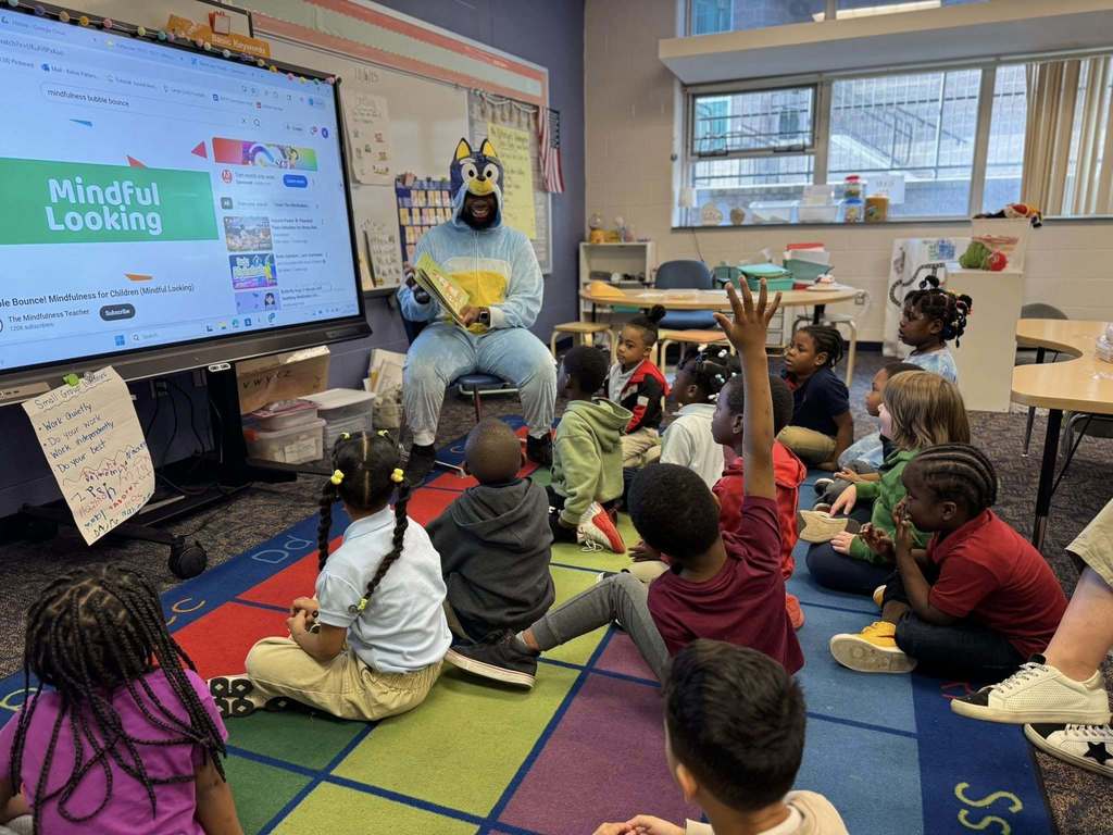 A district staff member is reading a storybook to a group of younger students who are seated on a colorful carper in a classroom. One of the students is raising his hand in the air.