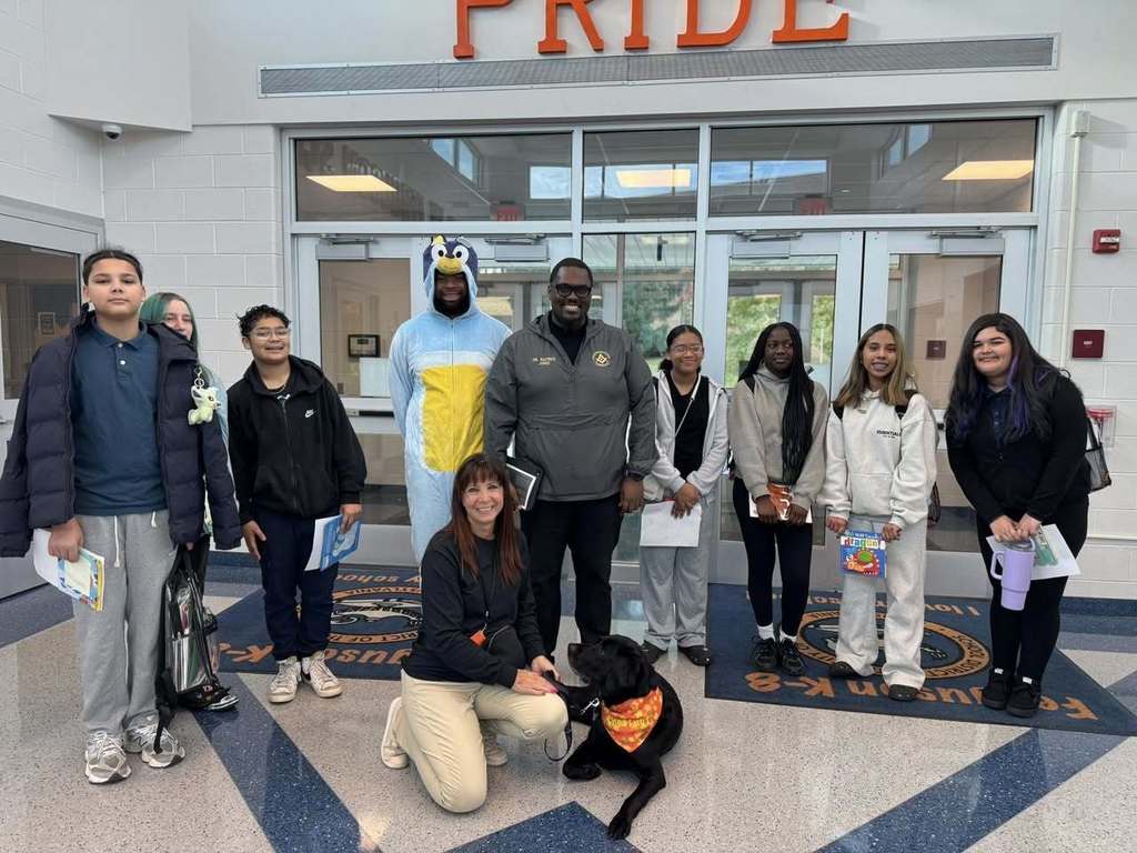 A group of district staff members and students are standing in a school lobby. A woman is kneeling beside a black dog who is seated. 
