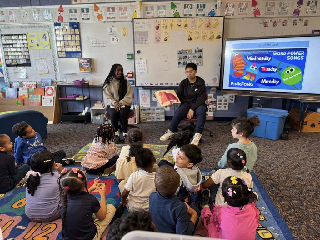 Two older students are reading a storybook to a group of younger students who are seated on a colorful carpet in a classroom. 