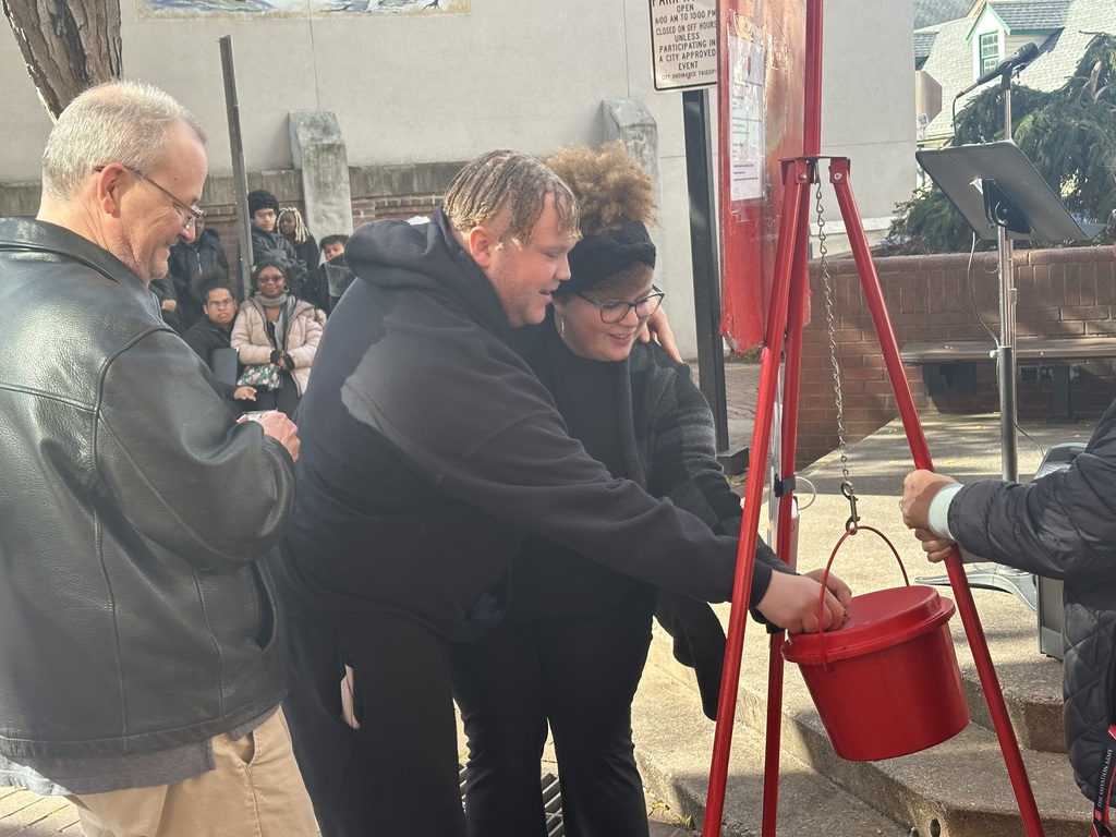 Two William Penn Senior High School students and a man are gathered around a red donation bucket outdoors. Both of the students are putting change into the bucket. Other students can be seen watching nearby in the background.