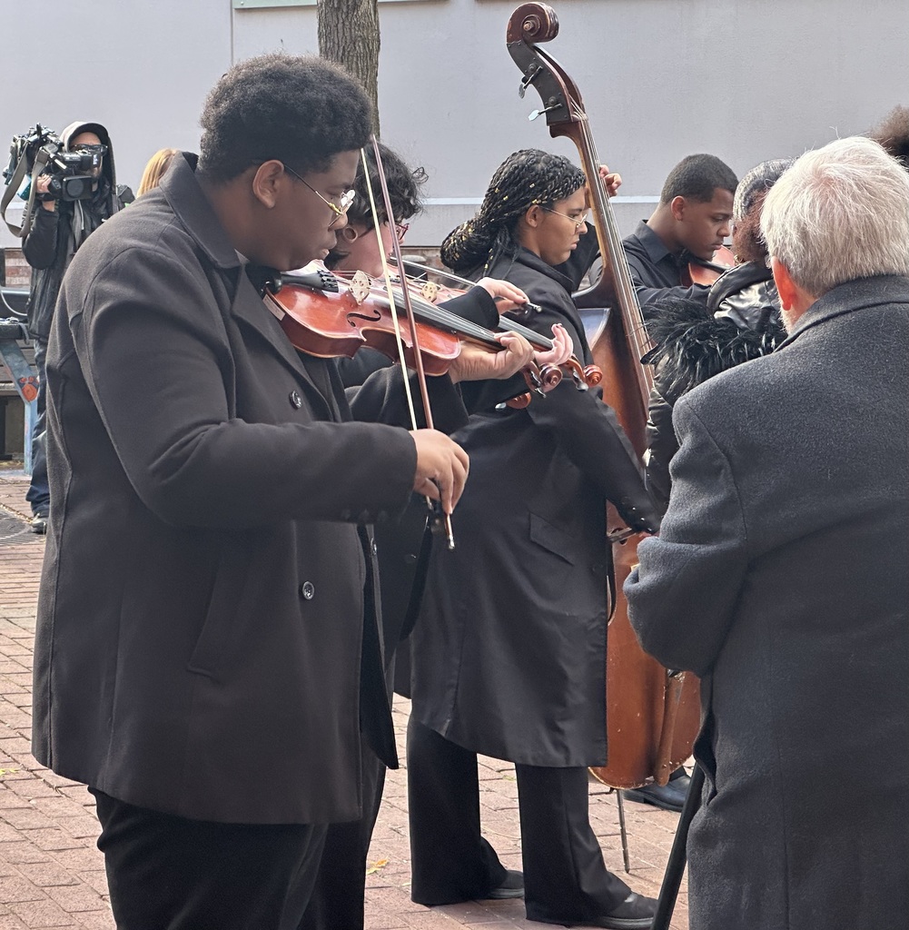William Penn Senior High School students are  performing a string ensemble outdoors, with violins and a double bass. A cameraman can be seen nearby filming the students.