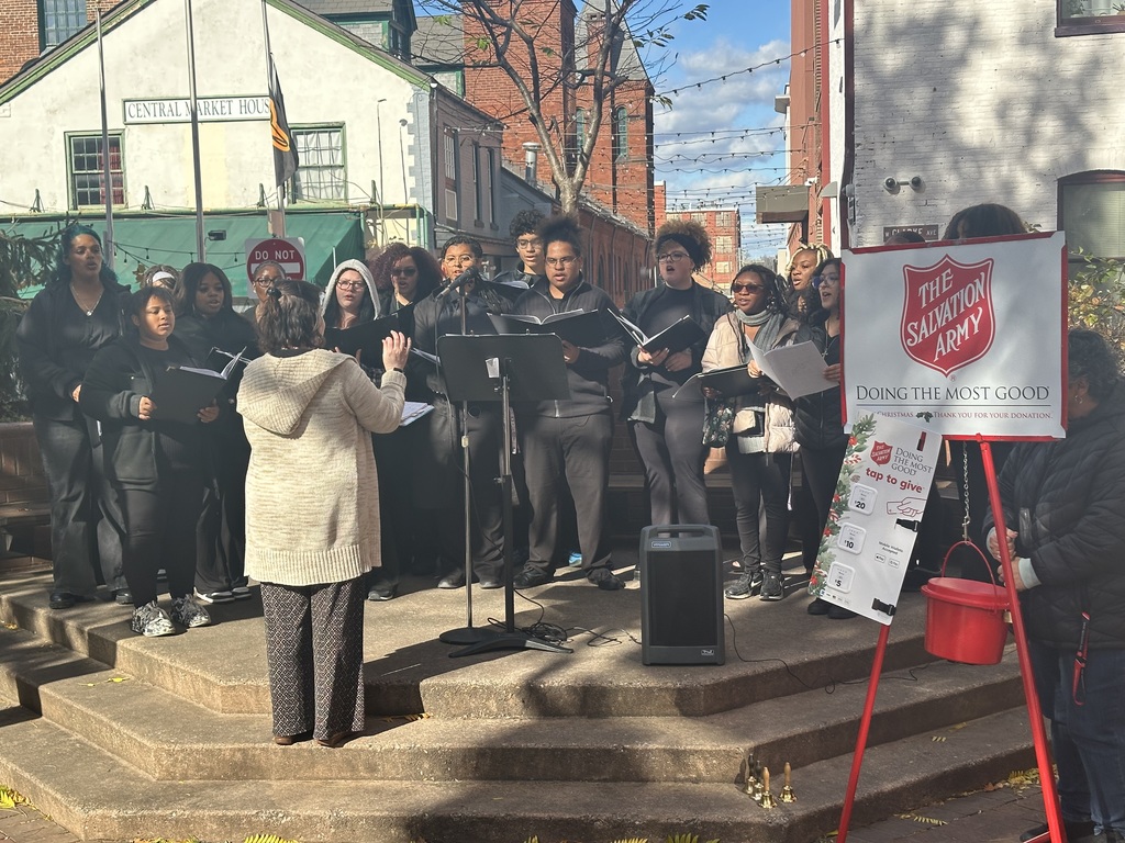 Students in the William Penn Senior High School choir are standing on a stage outdoors performing. A director is making gestures at them. A Salvation Army donation sign and red kettle can be seen nearby.