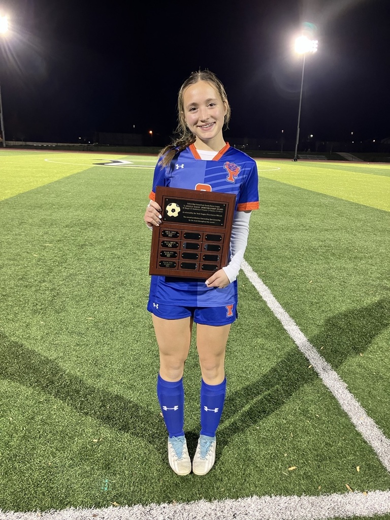 A York High girls soccer player is standing in the middle of a soccer field holding a plaque.