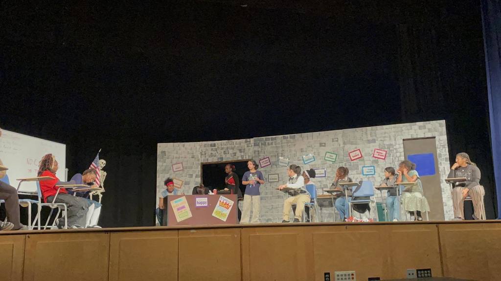 Students are performing a classroom play on stage featuring a brick wall backdrop, colorful posters, and desks. 
