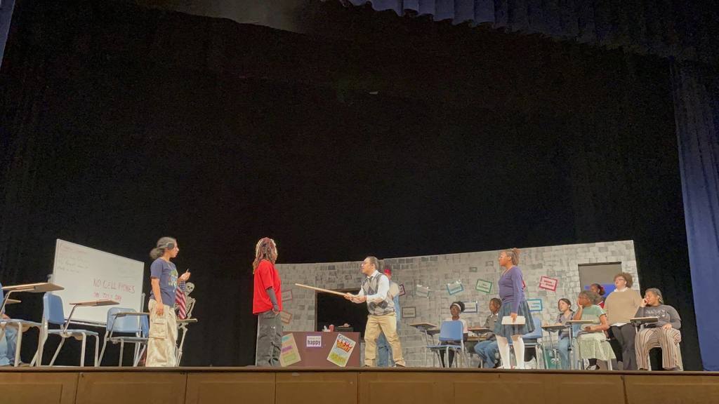 A school play scene on stage; a student is gesturing with a stick at another student. Other students are watching nearby, some of them standing up and others sitting at desks.