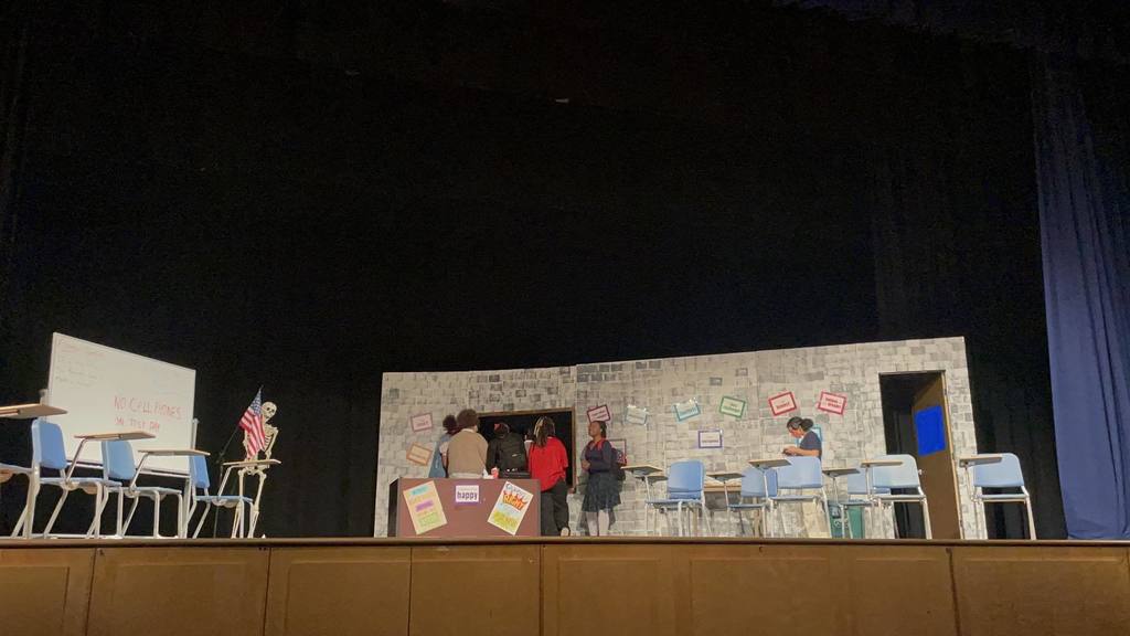 A stage set with a school classroom scene, featuring desks, chairs, a skeleton, a whiteboard, and an American flag. Several students are interacting with one another.