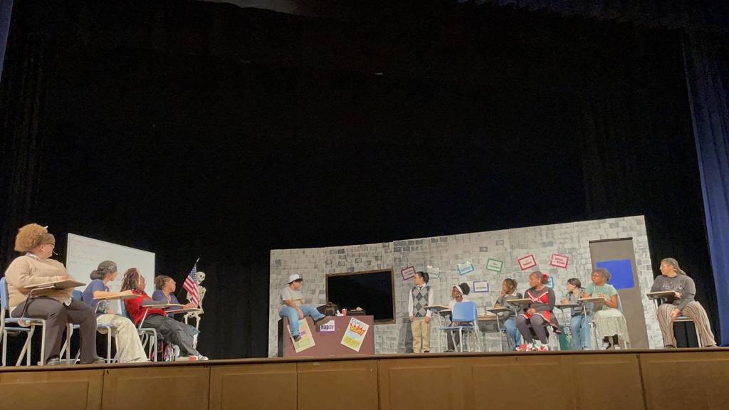 A school play scene with students sitting at desks, facing a teacher character at a decorated desk. The backdrop is a colorful classroom setting.