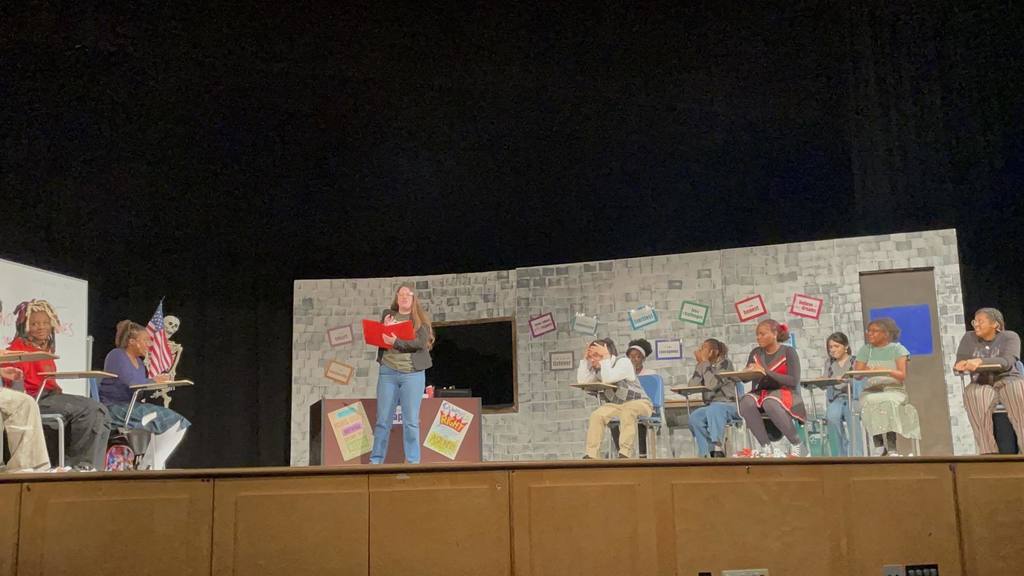 A group of students performing a school play on stage, set in a classroom backdrop. One student is standing reading a book, while other students are sitting at desks.