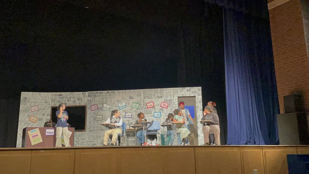 A group of students are performing on a theater stage set as a classroom. The backdrop features colorful words. 