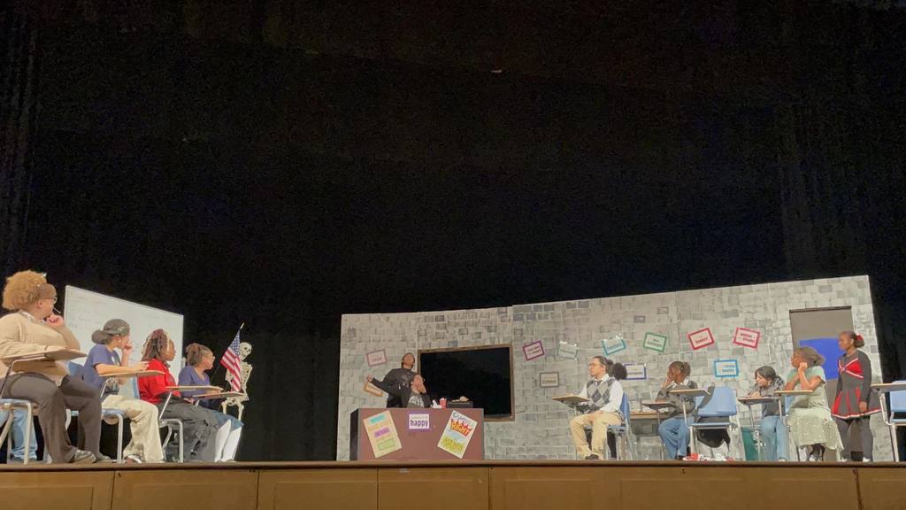 A school play depicting a classroom scene. Students are sitting at desks on both sides, with a teacher at the front. The backdrop shows a chalkboard, a skeleton, an American flag, and posters.