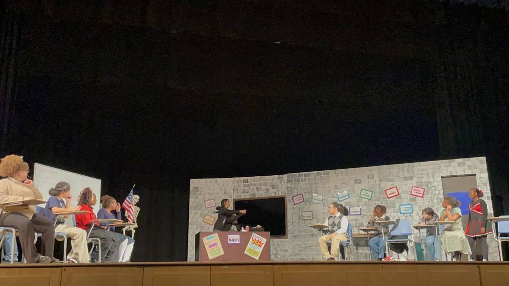 A school play set in a classroom with students seated at desks and a teacher at a podium. The backdrop is a brick wall with colorful posters.