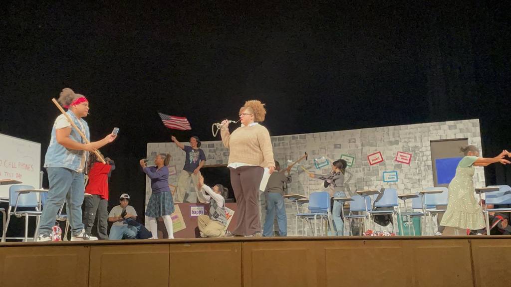 A lively stage scene with a group of students acting in a classroom setting, one student is holding a megaphone. Energetic gestures and an American flag can be seen.