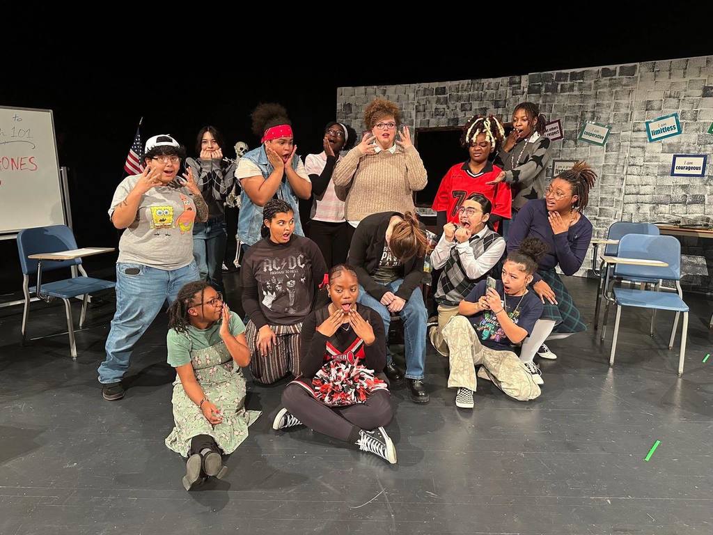 A group of students and a district staff member are in theatrical poses on stage, expressing surprise. They are surrounded by chairs and a brick wall backdrop with motivational signs.