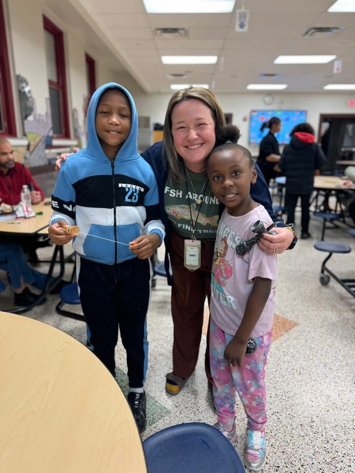 A district staff member and two students are standing in a school cafeteria near tables. Other people can be seen in the background. 