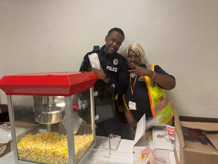 Two district staff members are standing behind a table that has a popcorn machine on it. One of the staff members is holding up the peace sign. 
