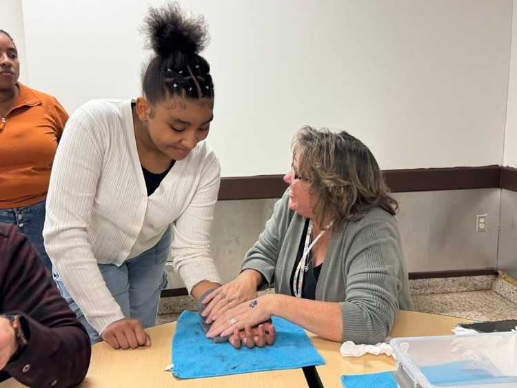 A district staff member is sitting at a table in a school cafeteria holding a rag on a students arm. Other people can be seen nearby. 