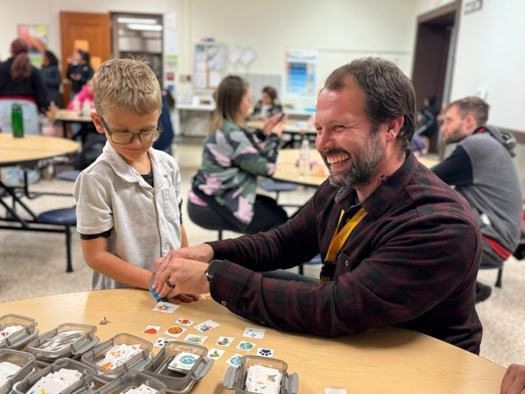 A district staff member is sitting at a table in a school cafeteria holding a rag on a students arm. Other people can be seen nearby. 