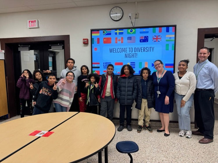 A group of district staff members and students are standing in front of a screen in a school cafeteria that says “Welcome to Diversity Night!”
