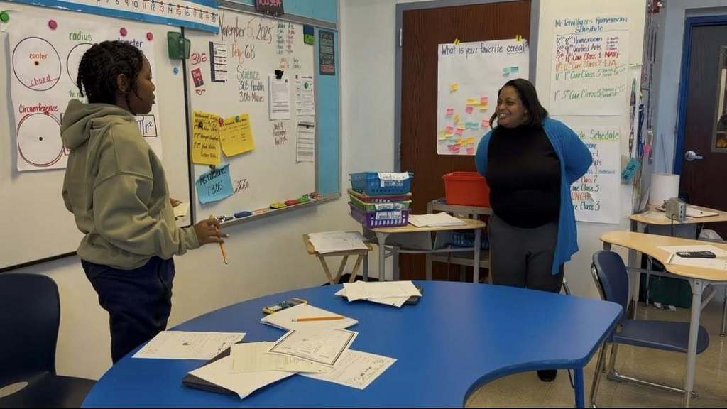Dr. Andrea Berry-Brown, Superintendent of Schools is standing near a blue table in a classroom engaging in a discussion with a student. There are posters that are hanging up on the walls.