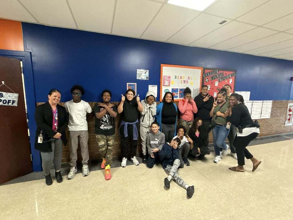 Dr. Andrea Berry-Brown, Superintendent of Schools, a group of students, and district staff members are posing in a school hallway. They are standing against a blue wall with bulletin boards, some of them holding up peace signs.