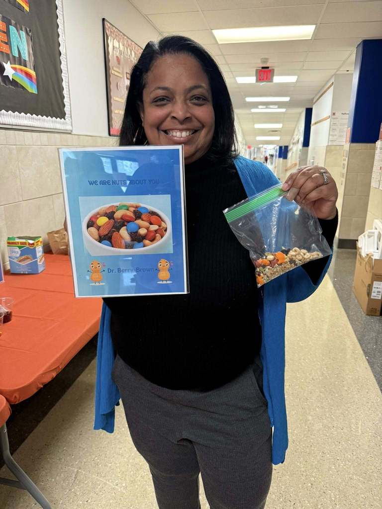 Dr. Andrea Berry-Brown, Superintendent of Schools is holding up a sign that says "We are nuts about you" and also a bag of mixed nuts, while standing in a school hallway.