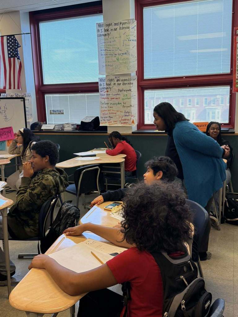 Dr. Andrea Berry-Brown, Superintendent of Schools is engaging with students in a classroom, surrounded by educational posters on the walls and an American flag hanging above.
