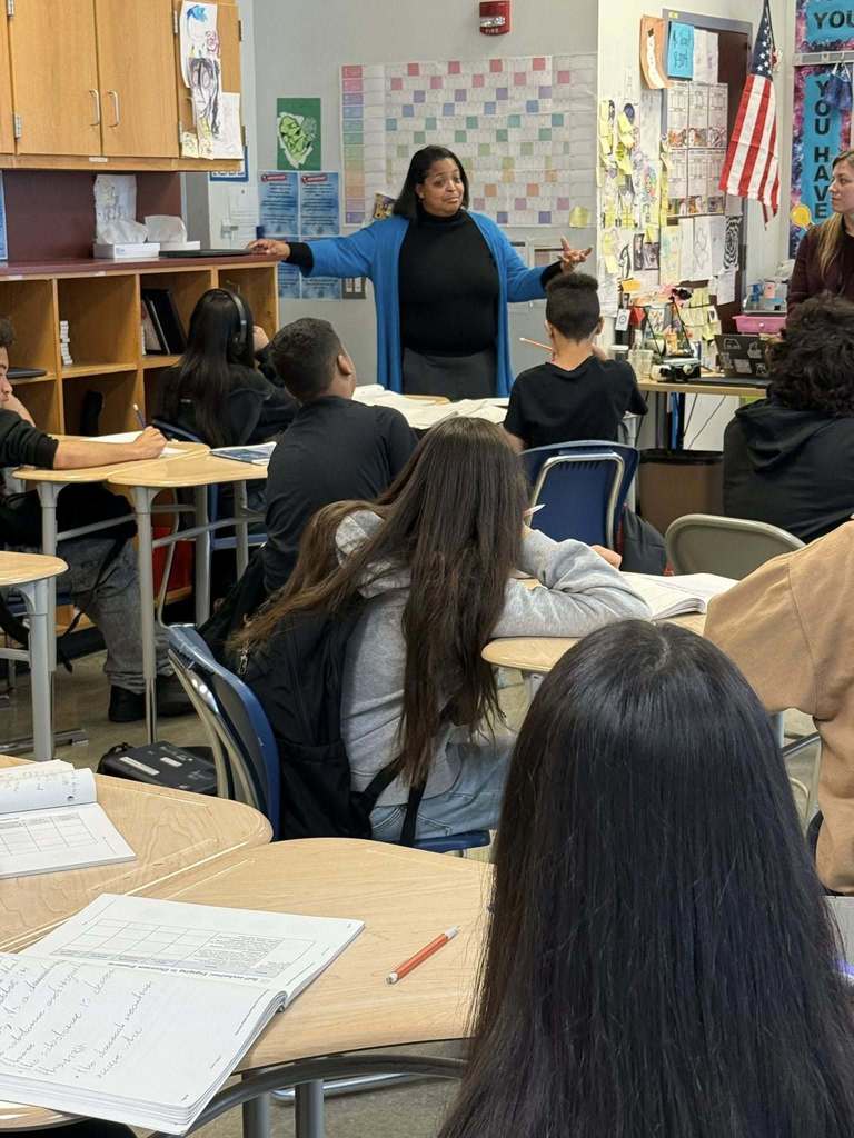 Dr. Andrea Berry-Brown, Superintendent of Schools is speaking to a class of students. The students are sitting attentively at their desks with open notebooks. The walls are filled with educational posters.