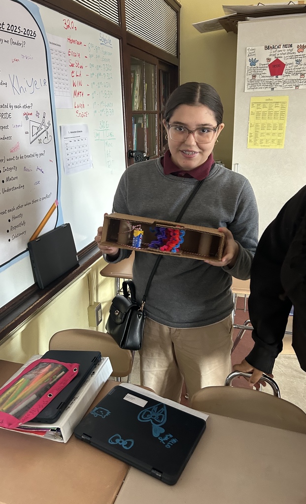 A student is holding a colorful craft project in a school classroom. A whiteboard with notes on it, a desk with two laptops and folders can be seen nearby.