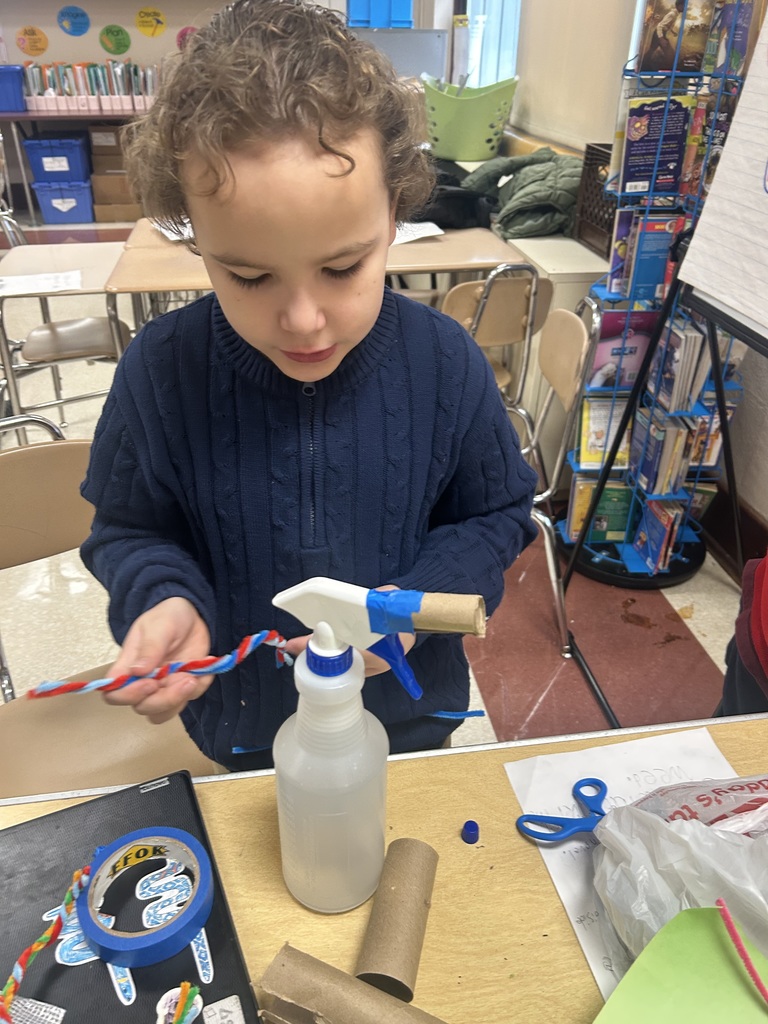 A student is working on a craft project in a school classroom using a spray bottle, colorful pipe cleaners, and cardboard. 