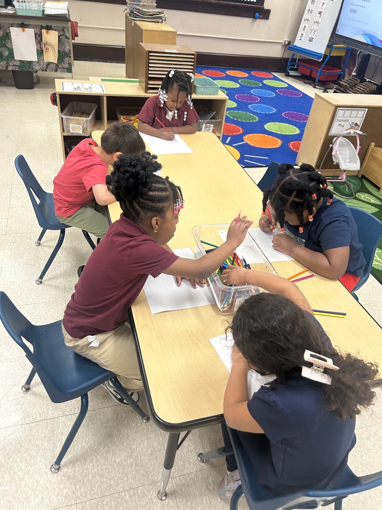 A group of five students are sitting around a table in a school classroom, drawing with crayons and colored pencils. 