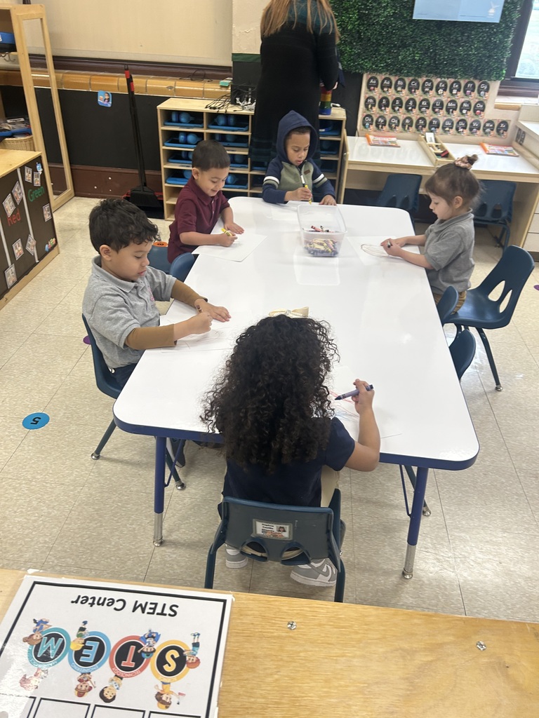 A group of five students are sitting at a table in a school classroom, coloring with crayons. A district staff member is standing nearby.