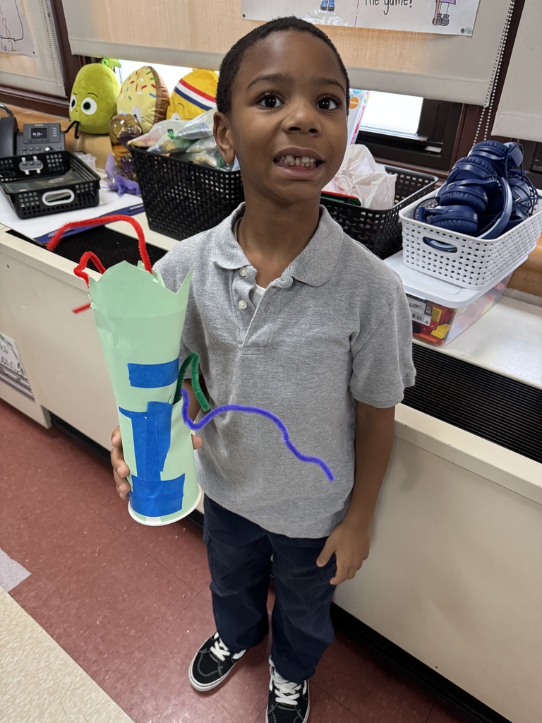 A student is holding a colorful handmade craft in a school classroom. The background shows baskets and craft supplies. 