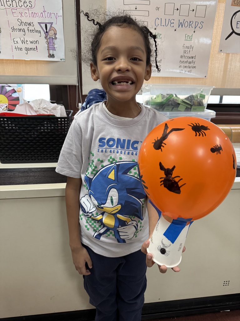 A student is holding an orange ballon with black bug drawings on it in a school classroom. Educational posters and baskets can be seen in the background.