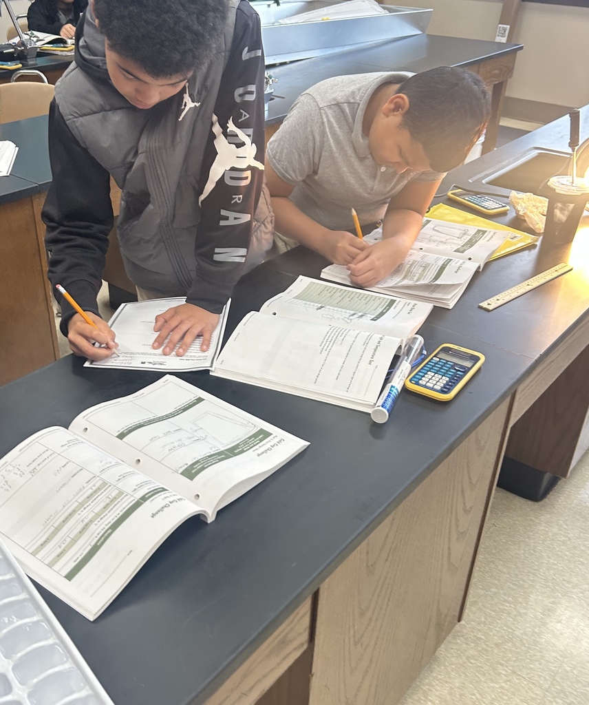 Two students are standing at desks in a school classroom writing on paper. A calculator can be seen nearby.