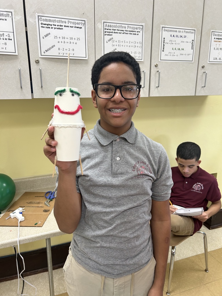 A student is holding a handmade stick puppet that is made out of cups in a school classroom that has educational posters. Another student can be seen sitting down reading in the background.