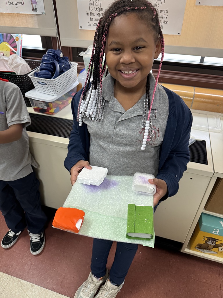 A student is holding a colorful foam craft project in a school classroom. The background shows another student as well as baskets, craft supplies and educational posters. 