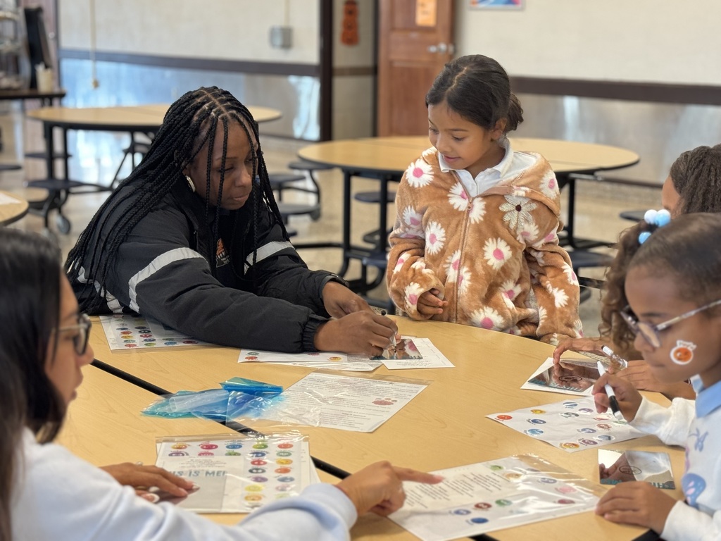 A district staff member and a group of students are seated at a table in a school cafeteria, engaging in an activity that involves stickers and mirrors.