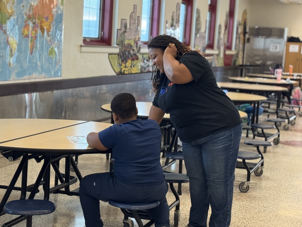 An adult is leaning over to assist a student who is seated at a table in a school cafeteria. A world map can be seen hanging on the wall in the background.