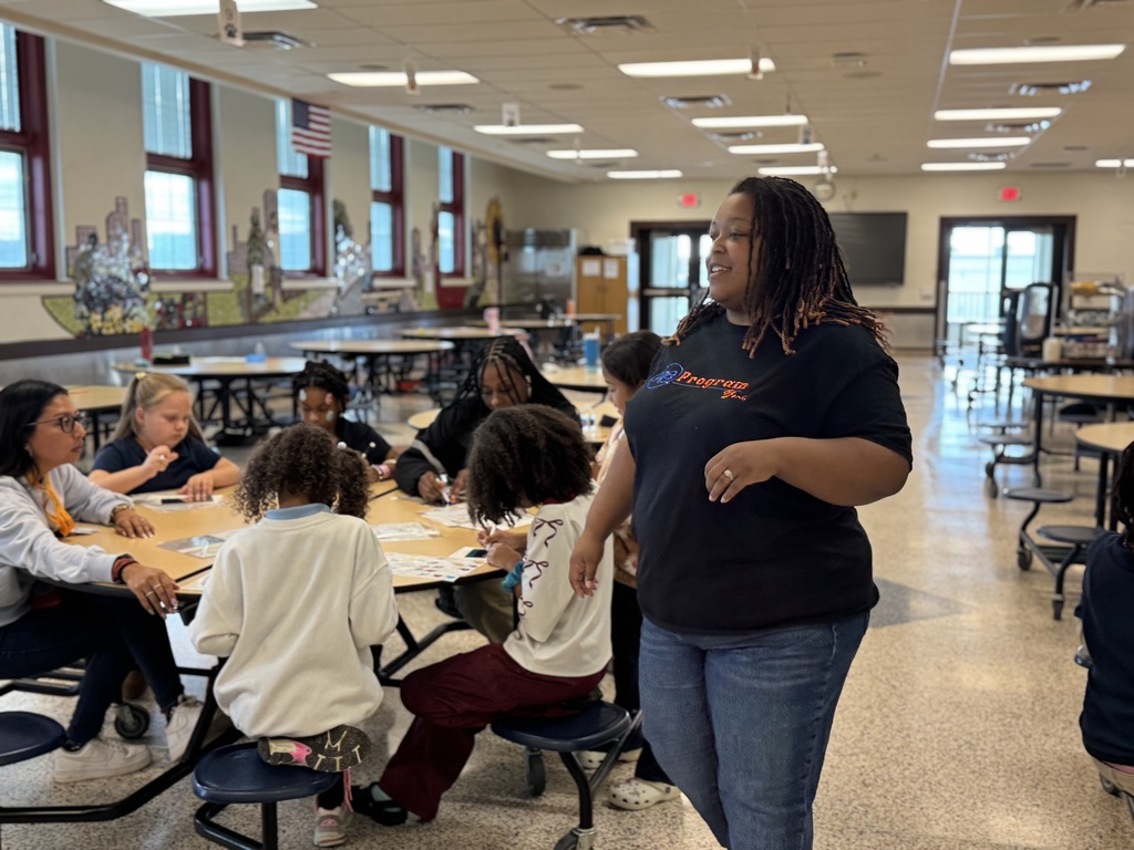 An adult is interacting with students who are seated at tables in a school cafeteria, engaging in an activity.