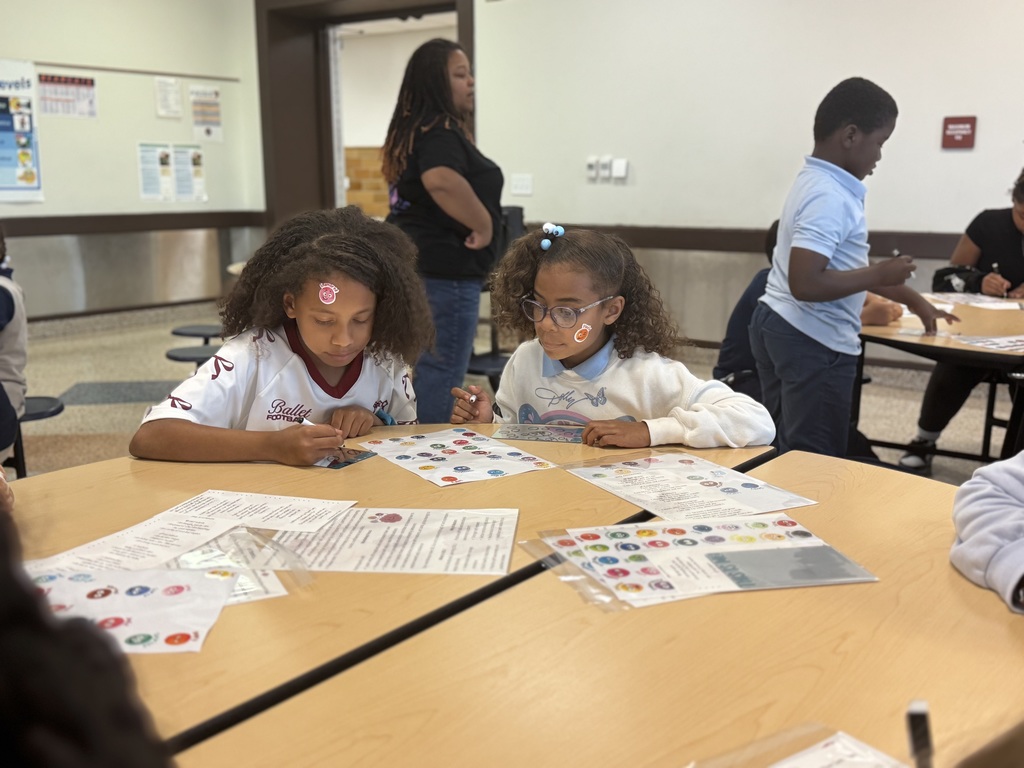 Two students with stickers on their faces are looking down at a piece of paper that has stickers on it. Other people can be seen in the background.