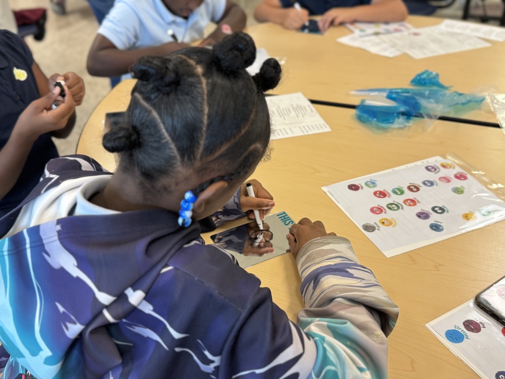A student is seated at a table in a school cafeteria engaging in an activity that involves stickers, markers, and mirrors.