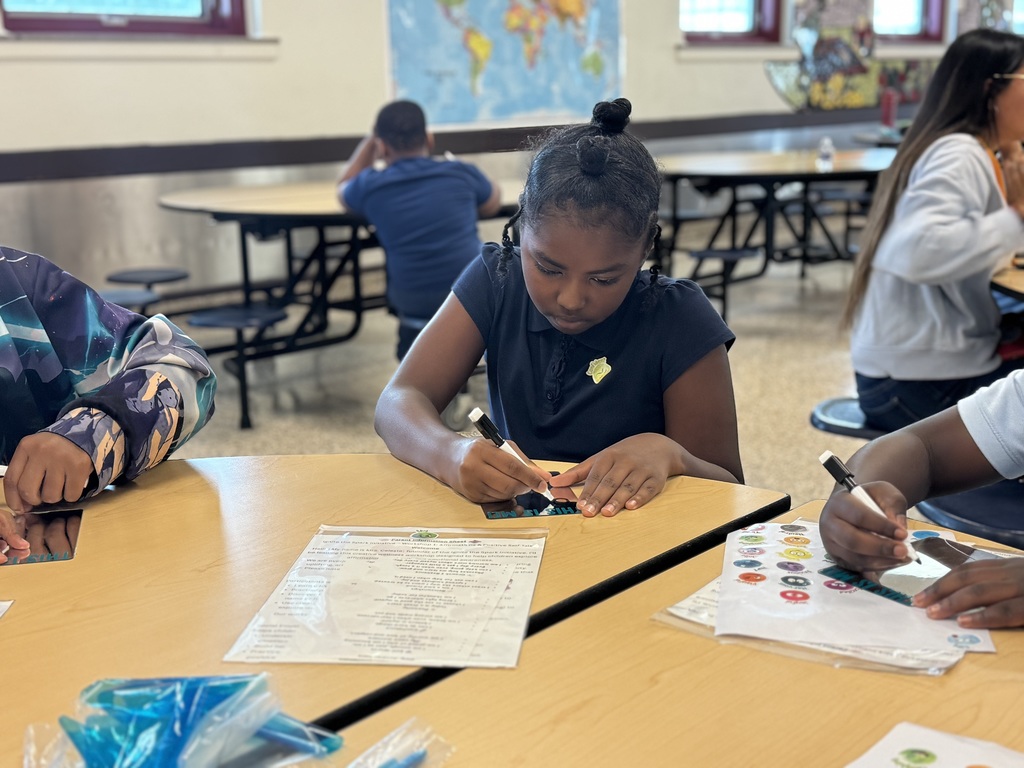 A student is sitting at a table in a school cafeteria drawing on a mirror with a marker. Other students can be seen sitting at tables nearby.