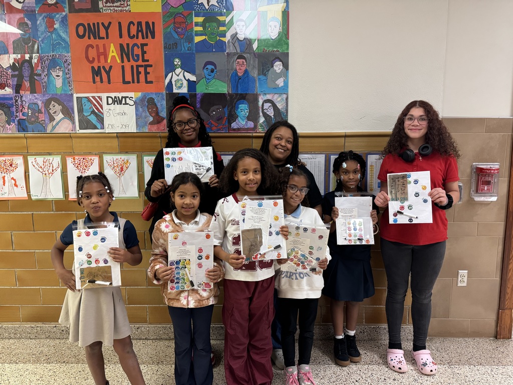 A group of students and one adult are standing in a school hallway holding up colorful art projects. The wall behind them is displaying vibrant artwork and a motivational quote.