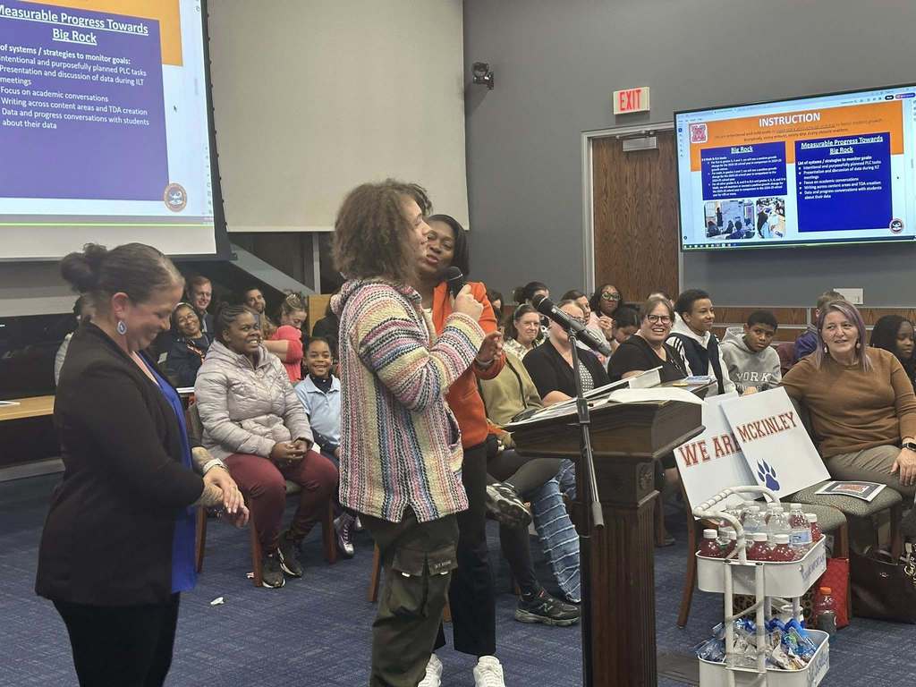 A male student is speaking into a microphone, held by a district staff member, at a podium in a conference room. There is an audience of people in the background looking attentively. District staff members are standing close to the student. A presentation is being displayed in the background.