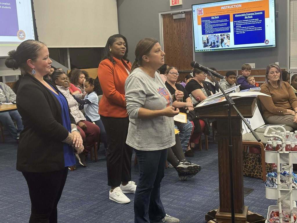 A district staff member is speaking into a microphone at a podium in a conference room. There is an audience of people in the background looking attentively. District staff members are standing close to the student. A presentation is being displayed in the background.