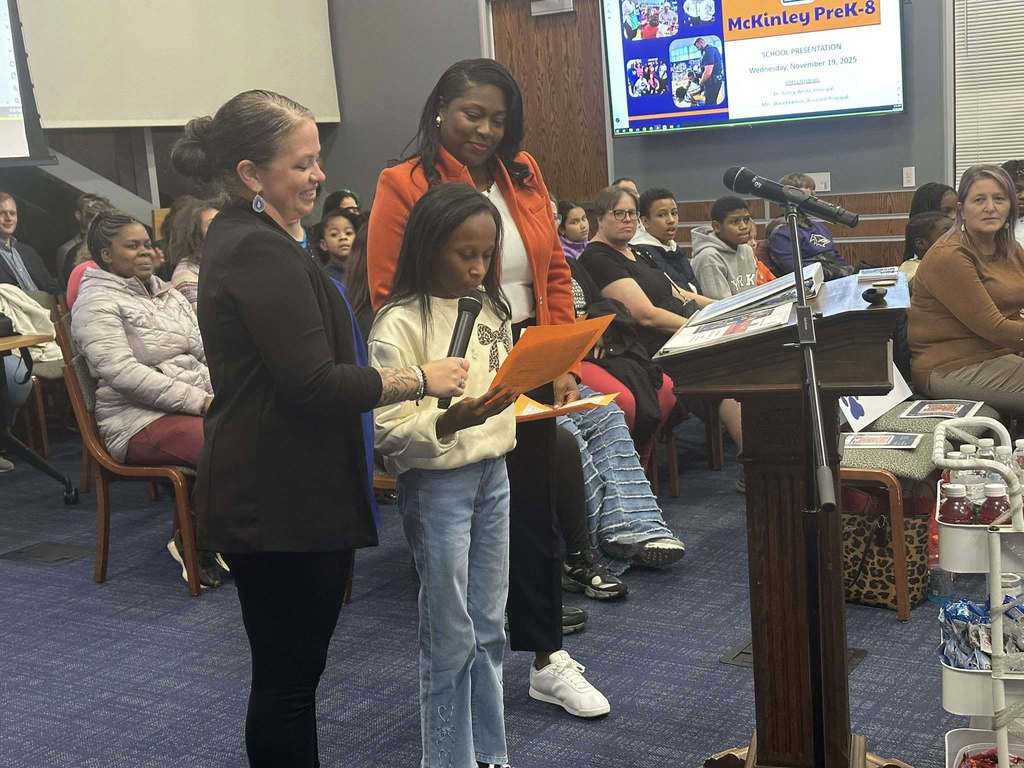 A female student is speaking into a microphone, held by a district staff member, at a podium in a conference room. There is an audience of people in the background looking attentively. District staff members are standing close to the student. A presentation is being displayed in the background.